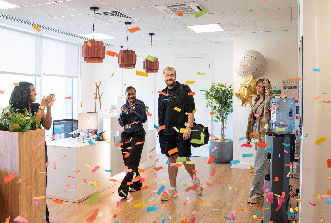 Liam walking into an office through falling confetti as Chaotic staff applaud, celebrating a £10,000 competition win