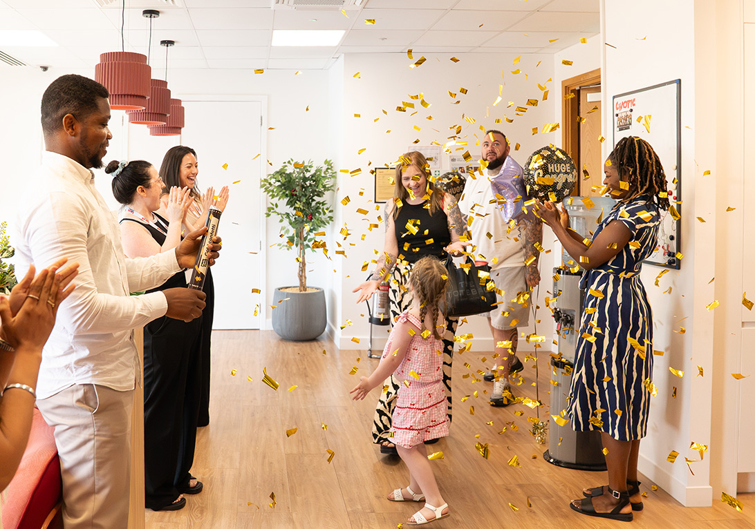 Heather walking into an office through falling confetti as Chaotic staff applaud, celebrating a £50,000 competition win