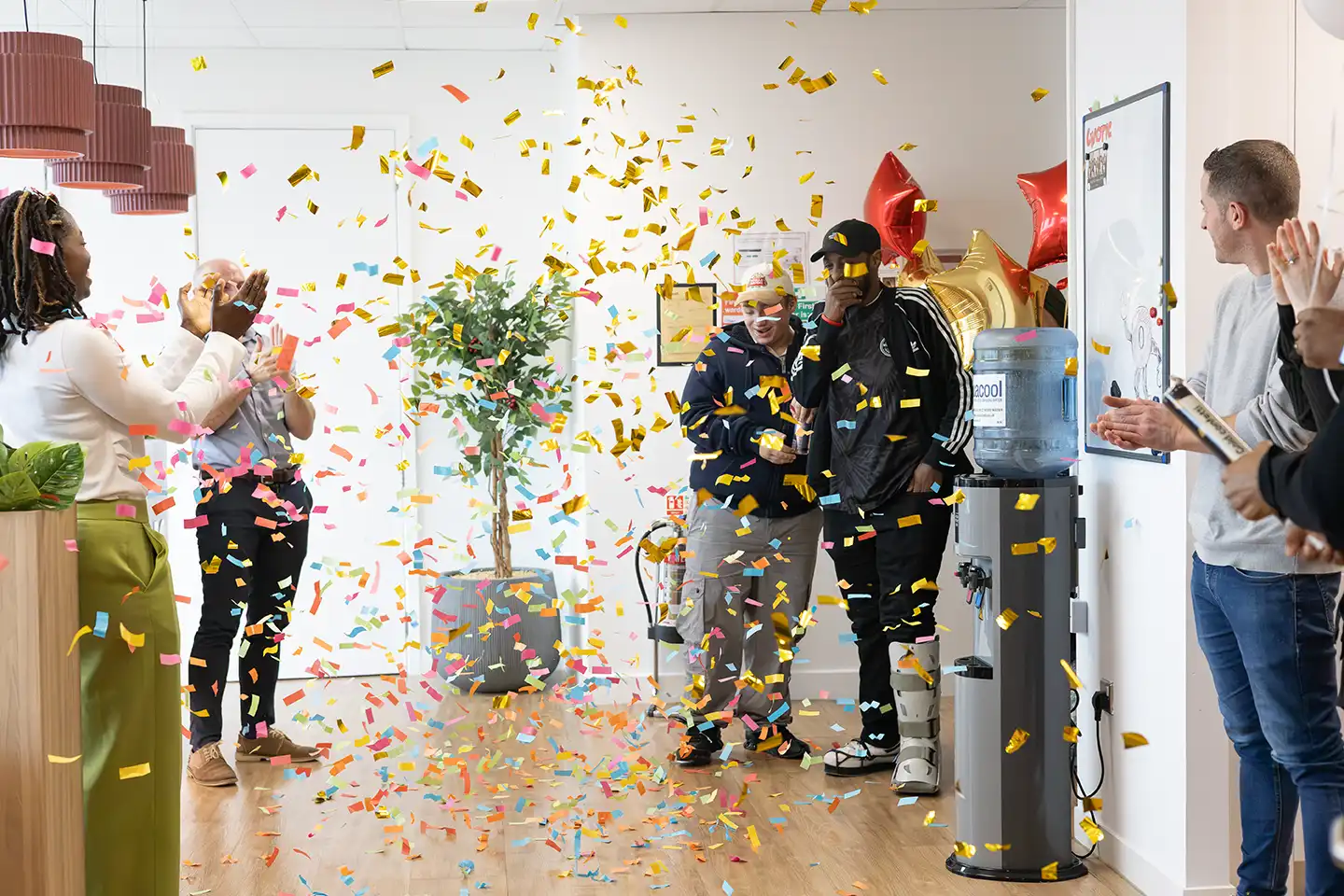 Khalid walking into an office through falling confetti as Chaotic staff applaud, celebrating a £40,000 competition win